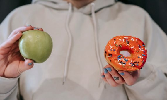 Crop unrecognizable female in loose hoodie showing healthy green apple and sweet delicious donut