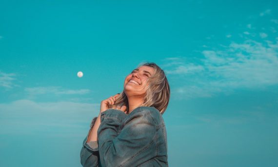 A happy woman in a denim jacket smiles under a vibrant blue sky with a visible moon.